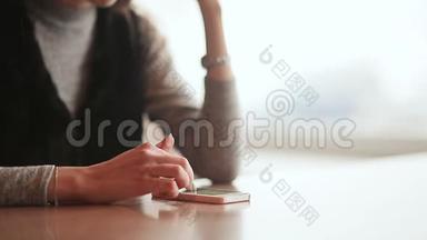 年轻漂亮的女孩手里拿着一部手机，坐在咖啡馆的桌子旁。 休息一下。 女孩的手`特写。